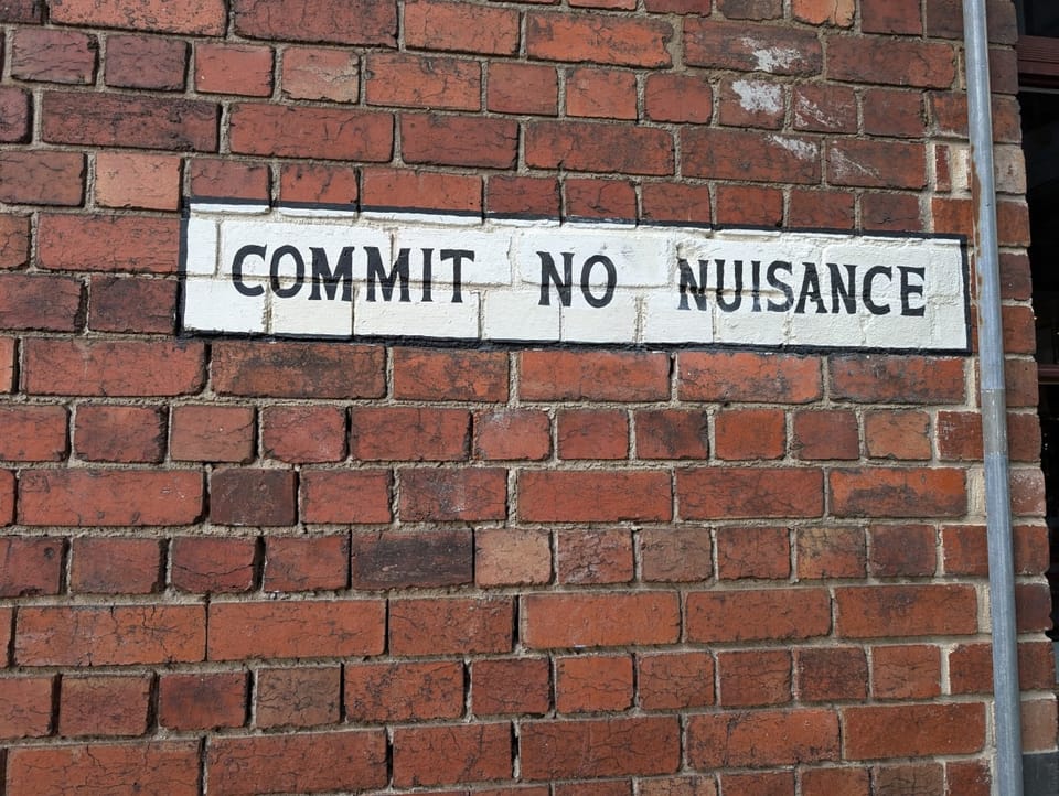 Photo of an old sign painted onto a red brick wall in black text on white paint saying "commit no nuisance"
