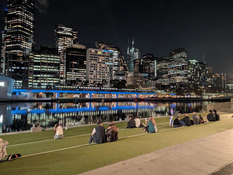 Photo of people sitting next to the Yarra River at Southbank at night with a view of the city skyline in front of them.